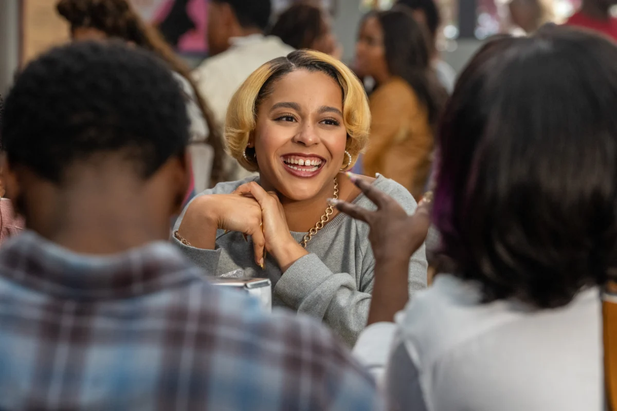 "Smiling woman with short blonde hair sits at a table, talking and laughing with friends in a lively, casual restaurant or café setting with other diners in the background."