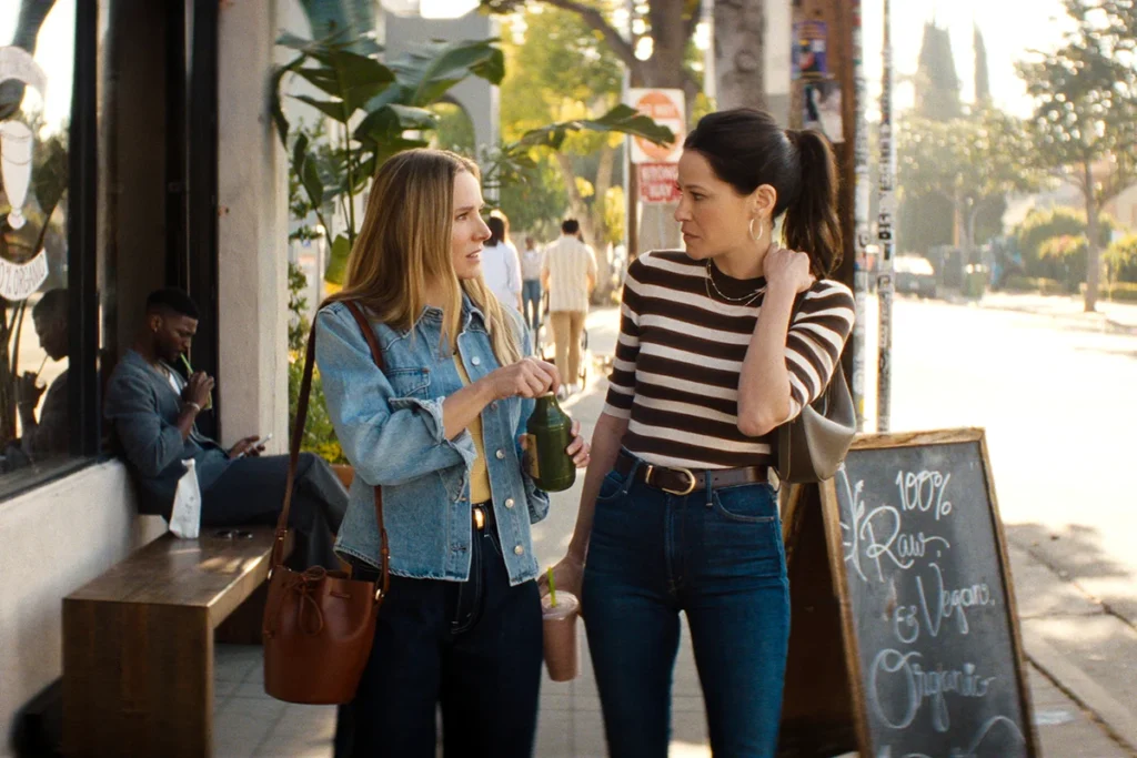 "Two women walk and talk on a sunny city sidewalk past a chalkboard sign and plants; one wears a striped shirt, the other a denim jacket. A man sits nearby on a bench with his phone and coffee. Urban outdoor setting."