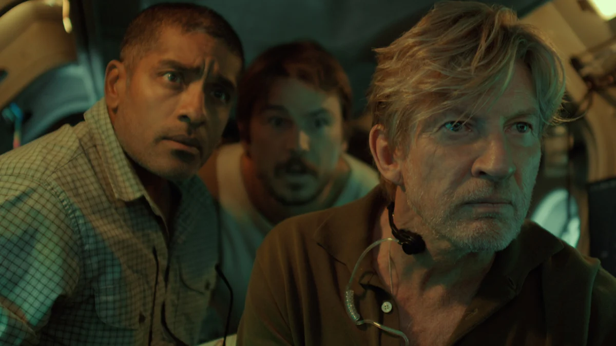 "Three men in a dimly lit submarine interior look intently at a screen, surrounded by equipment and controls, suggesting a tense or serious moment in an underwater or scientific environment."