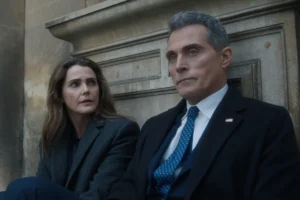 "Two people in formal coats sit on outdoor stone steps, appearing serious and deep in conversation, with an old stone wall in the background. The man wears a suit and tie with an American flag pin, suggesting a political or official context."